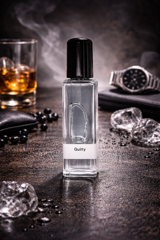 Perfume bottle labeled 'Guilty' on a dark surface with ice cubes, a glass of whiskey, and a watch.